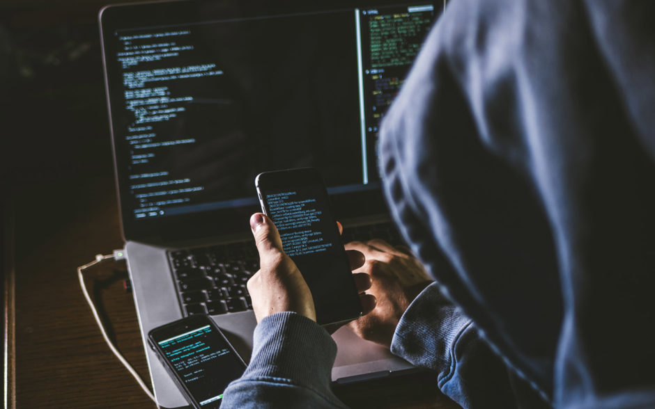 anonymous person in the hood sitting in front of computer working with laptop and mobile phone